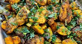 A tray of roasted chicken pieces and diced vegetables, including yellow courgette, onions, and herbs, garnished with chopped fresh parsley and spices. The dish appears golden-brown and flavourful.