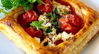 A golden puff pastry square filled with roasted cherry tomatoes, crumbled cheese, and herbs, served on a white plate with a side of fresh rocket.