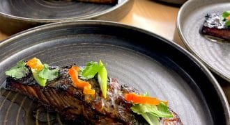 A close-up of a glazed salmon fillet garnished with sliced red peppers, spring onions, and coriander, served on a dark ceramic plate with more plated salmon portions in the background.