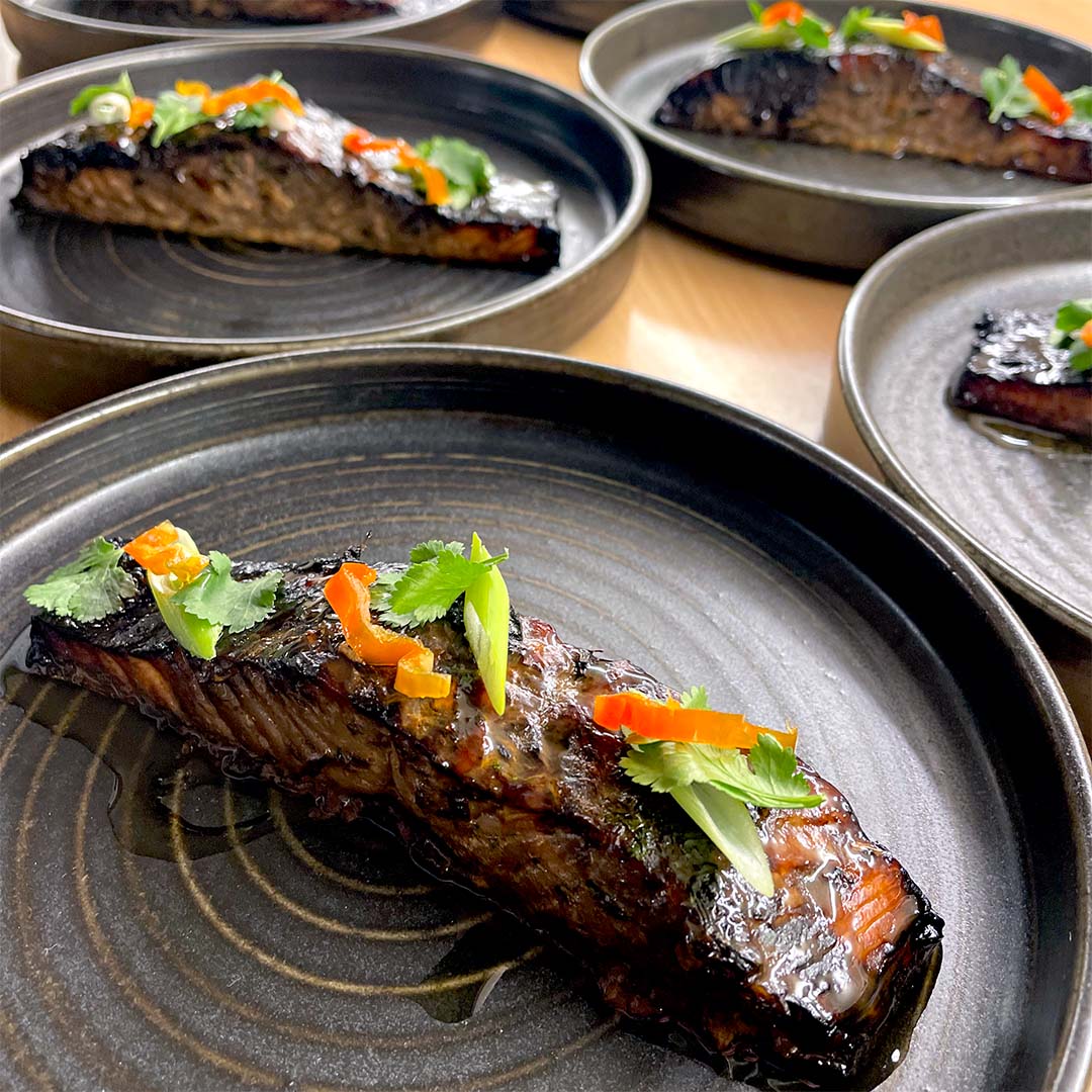A close-up of a glazed salmon fillet garnished with sliced red peppers, spring onions, and coriander, served on a dark ceramic plate with more plated salmon portions in the background.