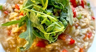 A close-up of creamy risotto with diced red peppers, garnished with a handful of fresh rocket, served in a white bowl.