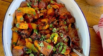 A white oval dish filled with a colourful roasted vegetable medley, including sweet potatoes, peppers, and mushrooms, garnished with chopped herbs. A spoon rests in the dish, and salt and chilli flakes are nearby.