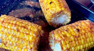 Three pieces of grilled corn on the cob, coated with seasoning and butter, sit on a black tray with melted butter and spices pooling around them.