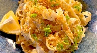 A bowl of fettuccine pasta topped with golden breadcrumbs and fresh herbs, garnished with fennel fronds and served with a lemon wedge on the side.