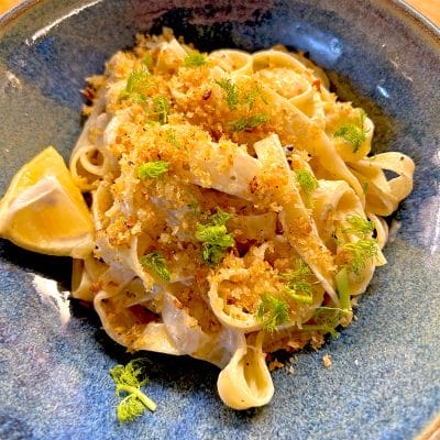 A bowl of fettuccine pasta topped with golden breadcrumbs and fresh herbs, garnished with fennel fronds and served with a lemon wedge on the side.