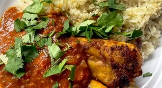 A plate of chicken curry with rich red sauce, served alongside seasoned rice and garnished with fresh coriander leaves.