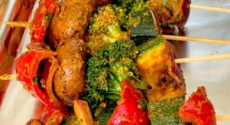 Vegetable skewers with mushrooms, broccoli, courgette, red peppers, and onions covered in seasoning, arranged on a foil-lined tray.