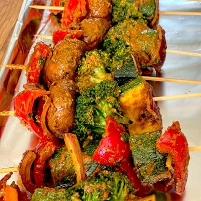 Vegetable skewers with mushrooms, broccoli, courgette, red peppers, and onions covered in seasoning, arranged on a foil-lined tray.