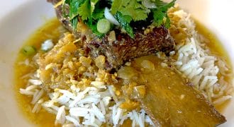 A braised beef short rib served over white rice, topped with fresh coriander and chopped spring onions, with a golden sauce poured around the dish on a white plate.