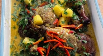 A roasting dish filled with roasted chicken pieces in a yellow curry sauce, garnished with pineapple chunks, chopped coriander, and sliced red chilli peppers.