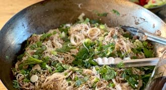 A large pan filled with cooked rice noodles, chopped spring onions, herbs, and sesame seeds, with metal tongs resting on the side. In the background, there is a bowl of mixed salad with tomatoes and cucumbers.