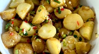A white bowl filled with halved baby potatoes mixed with chopped spring onions, red chilli, and herbs, placed on a wooden surface.