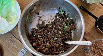 A metal pan with cooked minced meat and chopped spring onions sits on a wooden table, surrounded by lettuce leaves, a bowl of rice, cutlery, and small dishes with various toppings.