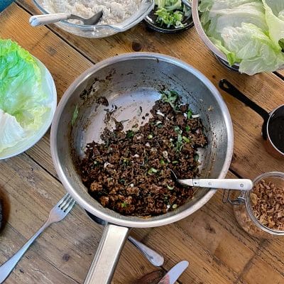 A metal pan with cooked minced meat and chopped spring onions sits on a wooden table, surrounded by lettuce leaves, a bowl of rice, cutlery, and small dishes with various toppings.
