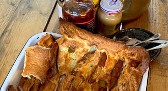 A tray of toad in the hole (sausages baked in Yorkshire pudding batter) sits on a wooden table next to a pot of mashed potato, jars of condiments, and some serving utensils.