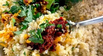A close-up of cooked rice mixed with fresh chopped herbs and a spoonful of red sauce, stirred together in a white bowl with a metal spoon.