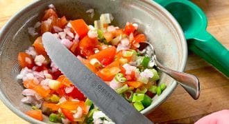 A hand holds a knife above a bowl of chopped tomatoes, onions, and green peppers. Nearby are a spoon, a green citrus squeezer, a bottle of white wine vinegar, and a bowl with a brown liquid.