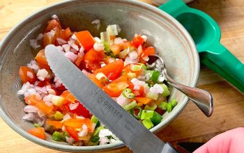 A hand holds a knife above a bowl of chopped tomatoes, onions, and green peppers. Nearby are a spoon, a green citrus squeezer, a bottle of white wine vinegar, and a bowl with a brown liquid.