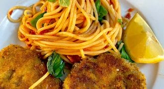 A plate with a serving of spaghetti in tomato sauce, garnished with basil leaves, two pieces of breaded fried meat, and a lemon wedge on the side.