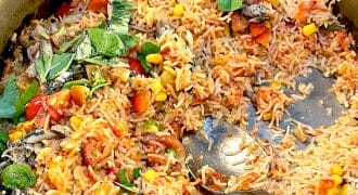 A large metal pan filled with mixed rice, sweetcorn, tomatoes, leafy herbs, and small pieces of fish. A serving spoon rests in the rice, which appears freshly cooked and partially served.