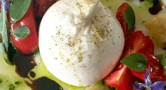 A plate with a ball of fresh burrata cheese, cherry tomato halves, toasted bread pieces, edible purple flowers, green leaves, olive oil, and a drizzle of balsamic glaze.