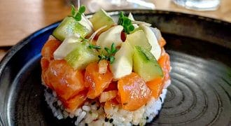 A round serving of sushi rice topped with diced salmon, avocado slices, diced cucumber, and microgreens, presented on a black plate with a glass of water in the background.