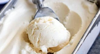 A metal ice cream scoop with a wooden handle is scooping vanilla ice cream from a rectangular metal container filled with smooth, creamy ice cream.