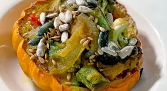A baked orange squash half filled with roasted vegetables, topped with sunflower, pumpkin, and sesame seeds, served on a white plate.