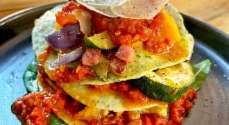 A stack of courgette slices layered with tomato sauce, diced vegetables, and pancetta, topped with grated cheese and black pepper, served on a dark plate with spinach leaves.