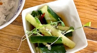 A small white bowl with cucumber slices, green herbs, and microgreens sits on a wooden table next to a bowl of white rice.