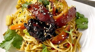 A white plate with stir-fried noodles, glazed vegetables, charred pieces of vegan mock duck, baby corn, carrots, fresh coriander leaves, and sesame seeds, with a fork resting on the edge.