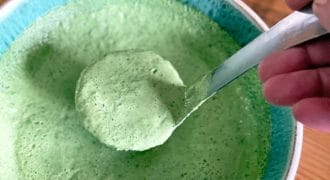 A hand holds a spoon scooping thick, green sauce or dressing from a turquoise-rimmed bowl on a wooden table. The sauce appears creamy and well-blended.