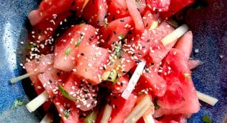 A bowl of watermelon pieces mixed with thinly sliced vegetables, topped with sesame seeds and chopped herbs, served in a blue ceramic dish.