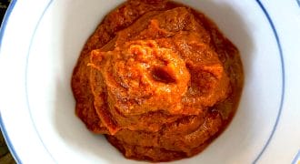 A white bowl with a blue rim filled with a thick, reddish-orange paste or sauce, placed on a wooden surface.