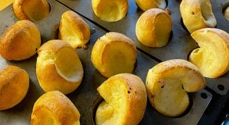 Golden-brown Yorkshire puddings rise and puff out of a black muffin tray, fresh from the oven, sitting on a wooden worktop.