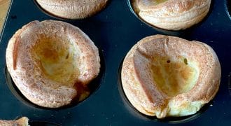 A close-up of freshly baked Yorkshire puddings in a muffin tray, showing their golden-brown, puffy edges and hollow centres.