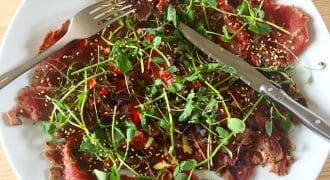 A plate of thinly sliced beef carpaccio topped with microgreens, sesame seeds, and chopped red peppers, with a fork and knife resting on the plate.