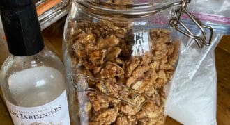 A large glass jar filled with candied walnuts sits on a wooden table next to a partially empty bottle of white wine. A plastic bag is in the background.