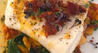 A fillet of white fish topped with microgreens and crispy bacon bits, served on a bed of wilted spinach, beans, and a tomato-based sauce on a white plate.