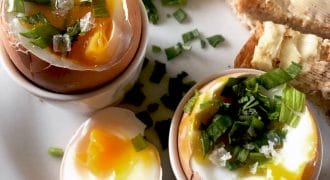 Two soft-boiled eggs in egg cups, topped with chopped spring onions and salt, with egg soldiers (buttered toast strips) and extra spring onions on a white plate. The yolks are runny and vibrant yellow.