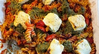 A baked pasta dish with spiral pasta, tomato sauce, dollops of green pesto, and several slices of melted white cheese on top, served in a white rectangular baking dish with a large serving spoon.