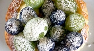 A pastry topped with green and purple grapes and dusted with icing sugar, placed on a white plate.