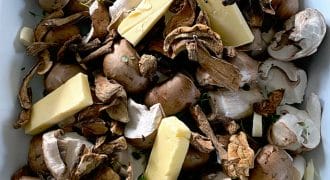 A close-up of raw mushrooms, both whole and sliced, mixed with dried mushrooms, sprigs of fresh herbs, and rectangular pieces of butter in a white dish.