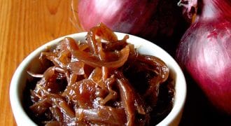 A small white bowl filled with caramelised onions sits on a wooden surface next to two whole red onions.