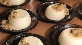 Several black plates, each with a serving of creamy white panna cotta topped with a small crumble and herbs, are arranged on a wooden surface, ready to be served.