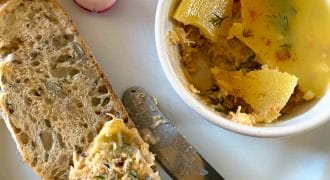 A slice of multigrain bread spread with a salmon pâté sits on a plate next to a bowl of pâté topped with jelly, a butter knife, and slices of fresh radish.