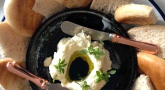 A black plate with a creamy white dip topped with olive oil and herbs, surrounded by slices of pitta bread, and two rose-gold handled spreading knives resting on the plate.