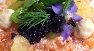 A colourful gourmet salad with orange segments, black caviar, citrus pieces, green herbs, edible flowers, and dollops of creamy sauce, artfully arranged on a white plate.