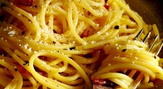 A close-up of cooked spaghetti on a plate, sprinkled with grated cheese and cracked black pepper, with a fork twirling some noodles.