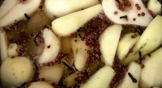 Sliced pears soaking in a saucepan with whole cloves, mustard seeds, and pink peppercorns, possibly being prepared for pickling or poaching.